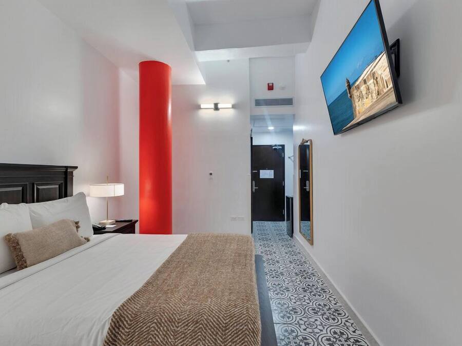 Habitación de hotel con cama, mesita de noche, lámpara, un pilar rojo, televisor montado en la pared y suelo estampado.