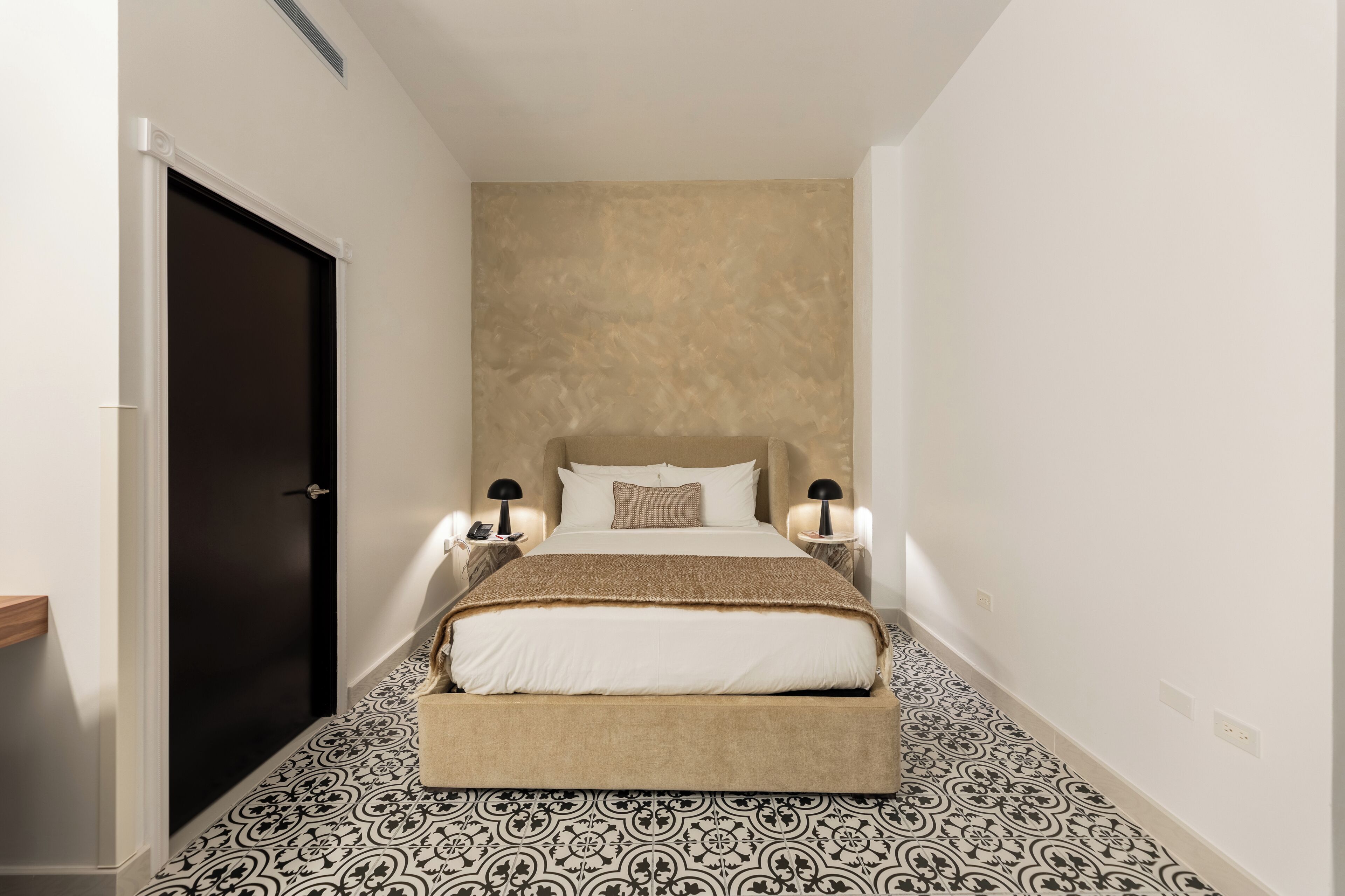 Un pasillo estrecho que conduce a una cama con cabecera, dos mesitas de noche con lámparas y baldosas con diseño en el suelo.