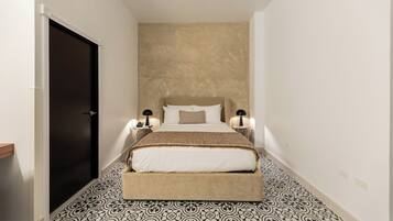 Un pasillo estrecho que conduce a una cama con cabecera, dos mesitas de noche con lámparas y baldosas con diseño en el suelo.