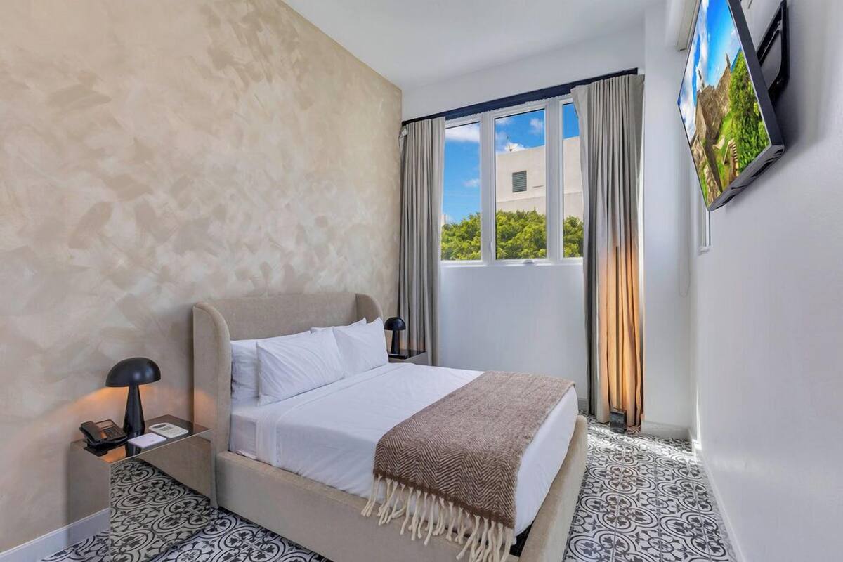 A hotel room with a bed, a nightstand with a lamp, a TV mounted on the wall, and a window with curtains.