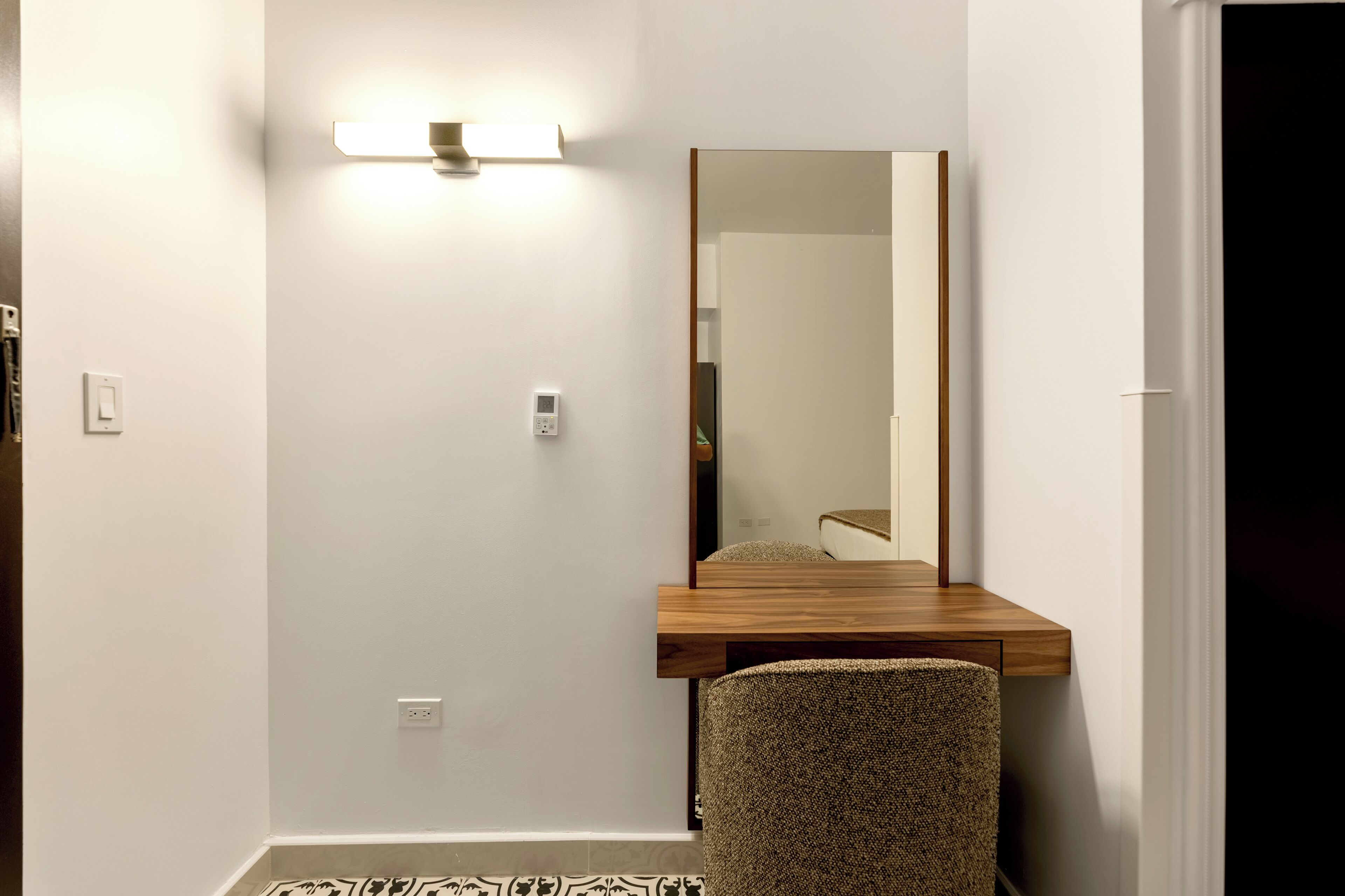 Une petite pièce avec un bureau en bois, une chaise, une lampe fixée au mur et un miroir.