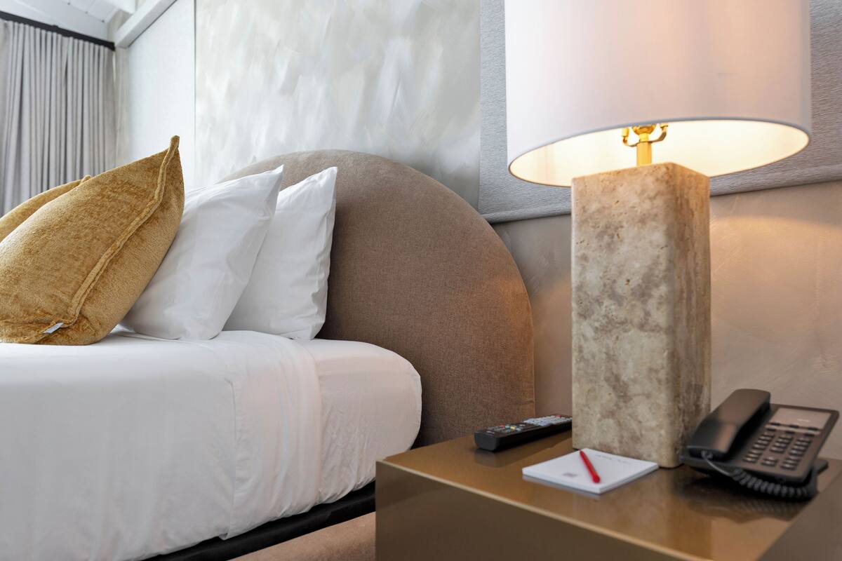 A hotel room with a bed, a nightstand with a lamp, a telephone, and a remote control.