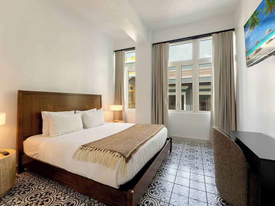 A hotel room with a wooden bed, two bedside tables with lamps, a large window with curtains, and a painting of a beach scene.