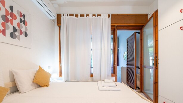 Una camera da letto con un letto, tende bianche, una porta in legno e vista sull'esterno.