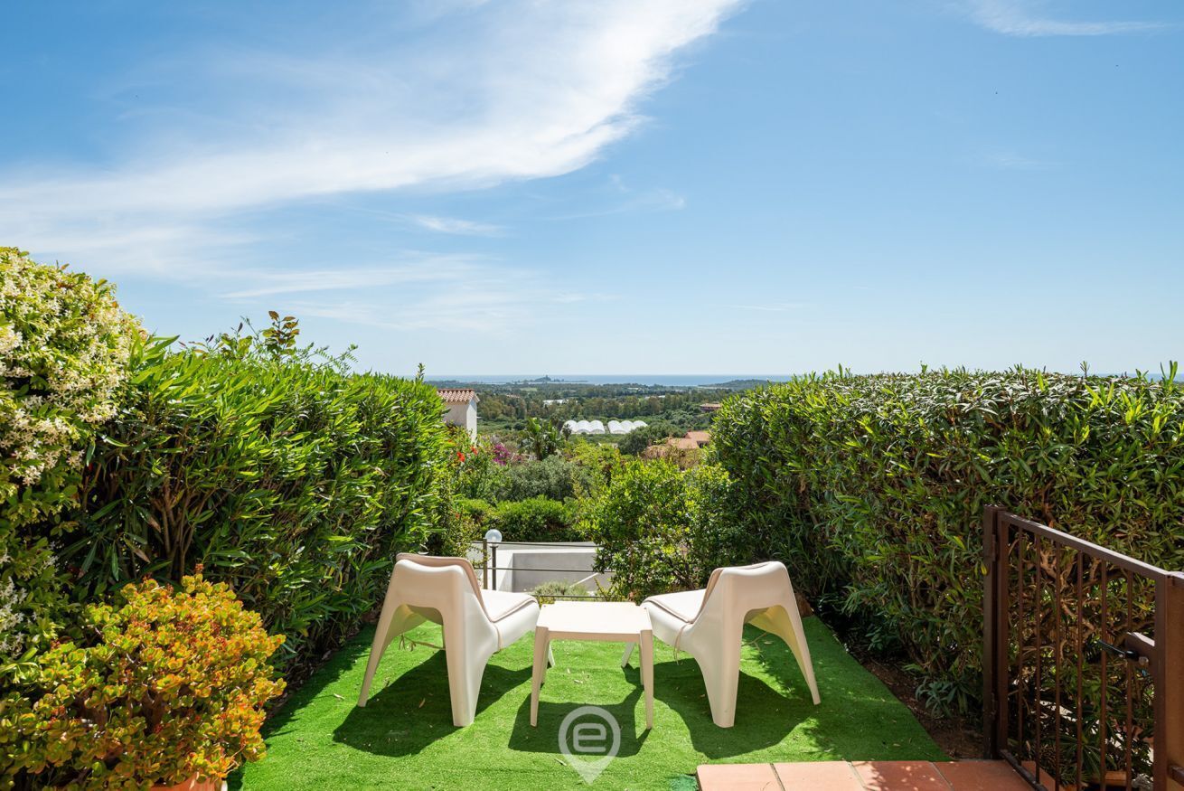 A patio with two white plastic chairs and a small table, surrounded by lush green hedges and a view of distant buildings.