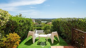Un patio con due sedie di plastica bianca e un tavolino, immerso in siepi verdi rigogliose e affacciato su edifici in lontananza.