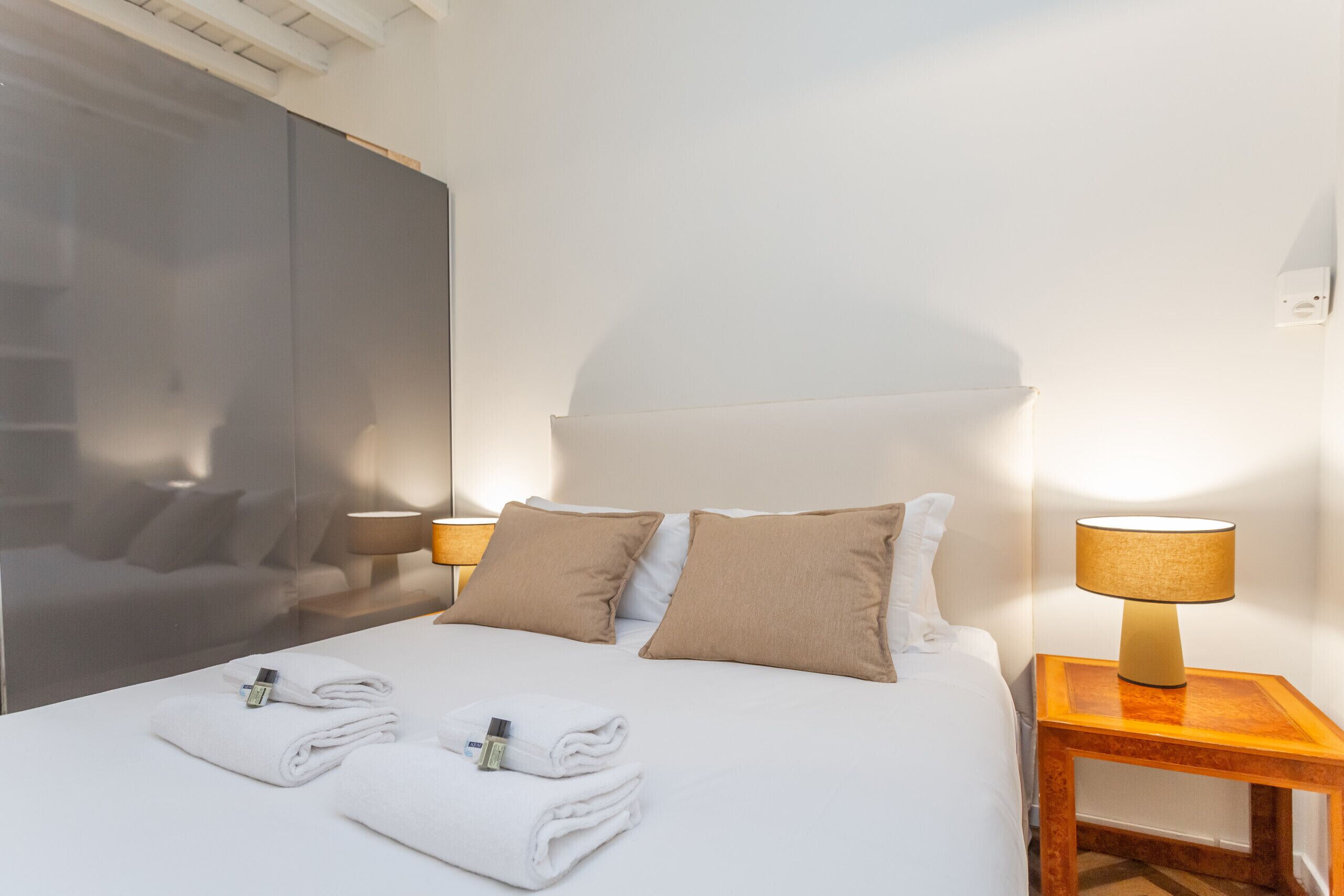 A neatly made bed with two pillows, a bedside table with a lamp, and a wall-mounted shelf.