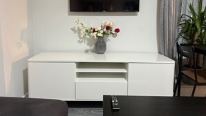 Een wit entertainment meubel met een flatscreen televisie erboven, een vaas met bloemen en een afstandsbediening op een tafel ernaast.