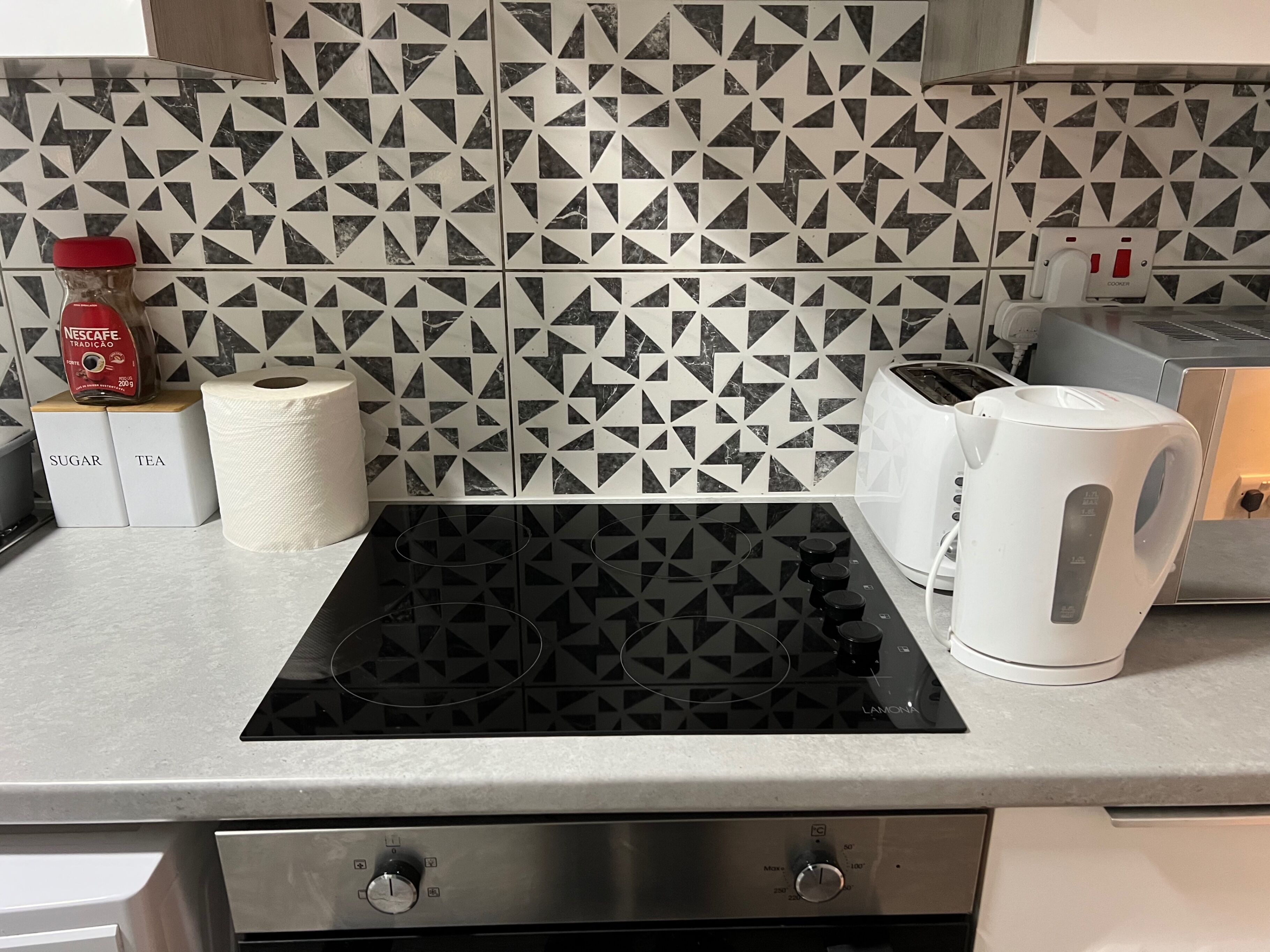 Uma cozinha com painel geométrico preto e branco, uma cafeteira Nescafé, um rolo de papel toalha, uma torradeira e uma chaleira.