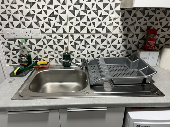 Una cocina con un fregadero, un escurreplatos, una botella verde y una caja etiquetada 'SUGAR' y 'TEA'.