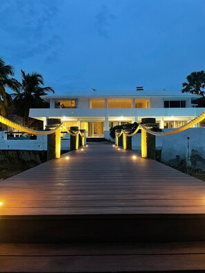 Un sendero de madera conduce a un edificio moderno de dos pisos con balcón, decorado con luces y cuerdas.