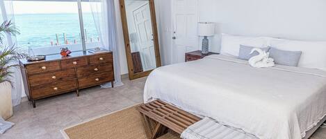 A bedroom with a large bed, a wooden bench, a dresser, and a view of the sea through a window.