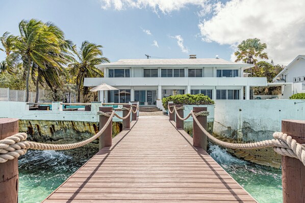 Un muelle de madera que lleva a un moderno edificio blanco con grandes ventanales, rodeado de palmeras y un cielo despejado.