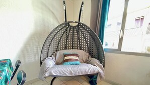 Ein Zimmer mit einem Hängestuhl, der eine gemusterte Rückenlehne, ein Kissen und ein Kopfkissen hat. Eine Gitarre lehnt an der Wand und ein Fenster mit Vorhängen ist zu sehen.