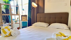 Ein ordentlich bezogenes Bett mit weißen Leinentüchern und zwei Kissen, eine Nachttischlampe sowie ein Regal mit Pflanzen und Dekoration.