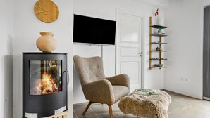 Un salon avec un poêle à bois, un fauteuil, un pouf rond, une horloge murale et un téléviseur fixé au mur.