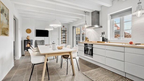 Une cuisine moderne avec des meubles blancs, un coin repas doté d’une table en bois et de chaises blanches, et une grande fenêtre qui laisse entrer la lumière naturelle.