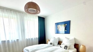 Een moderne slaapkamer met een houten bed, witte lakens, een geweven plafondlamp, een blauw kunstwerk en een nachtkastje met een lamp.
