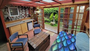 Un espace cuisine extérieur abrité, avec des murs en pierre, un évier et un réfrigérateur. On y trouve des chaises et une table en bois.