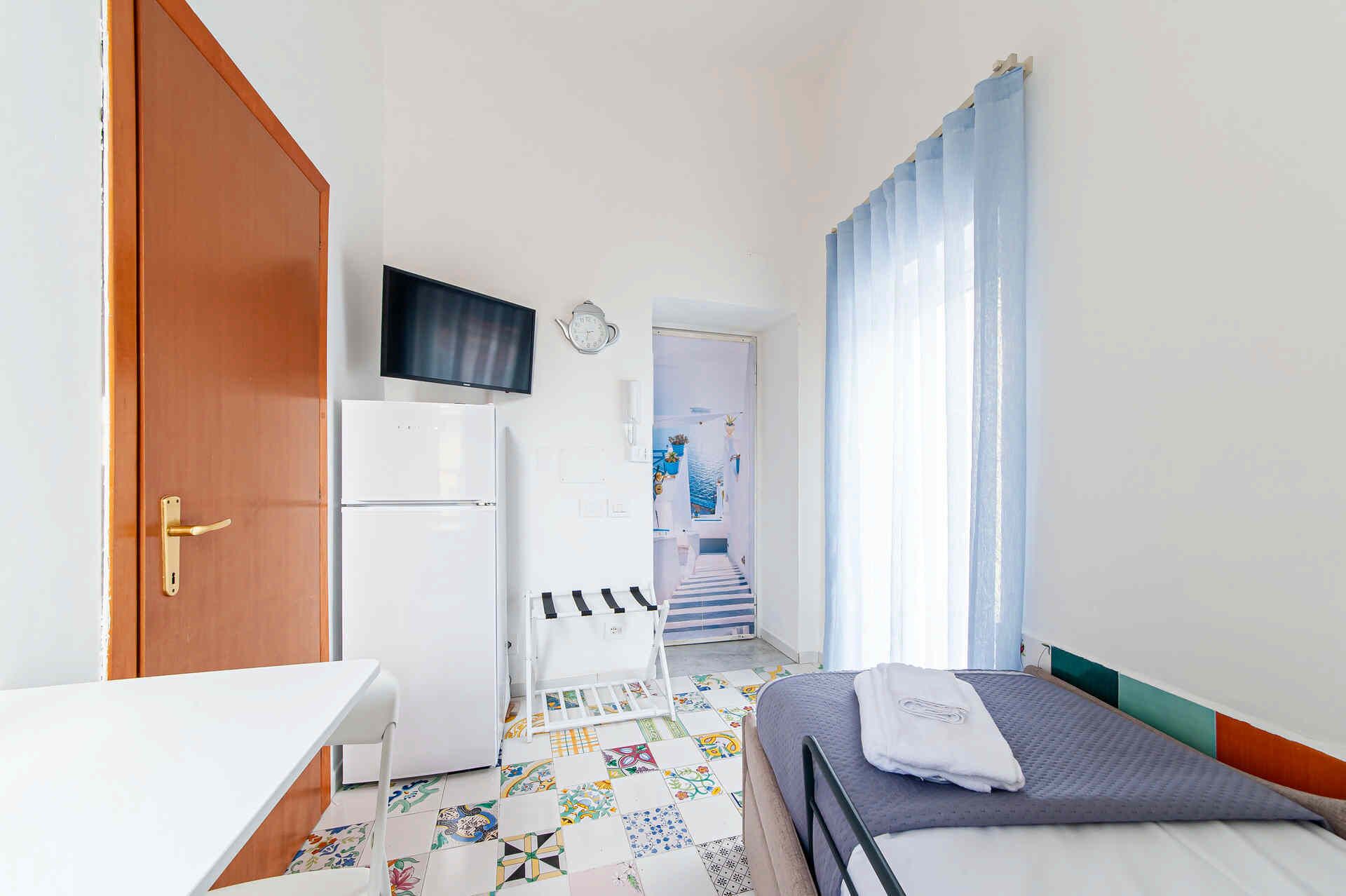 A compact hotel room with a bed, a desk, a refrigerator, a television, and a bathroom visible through an open door.