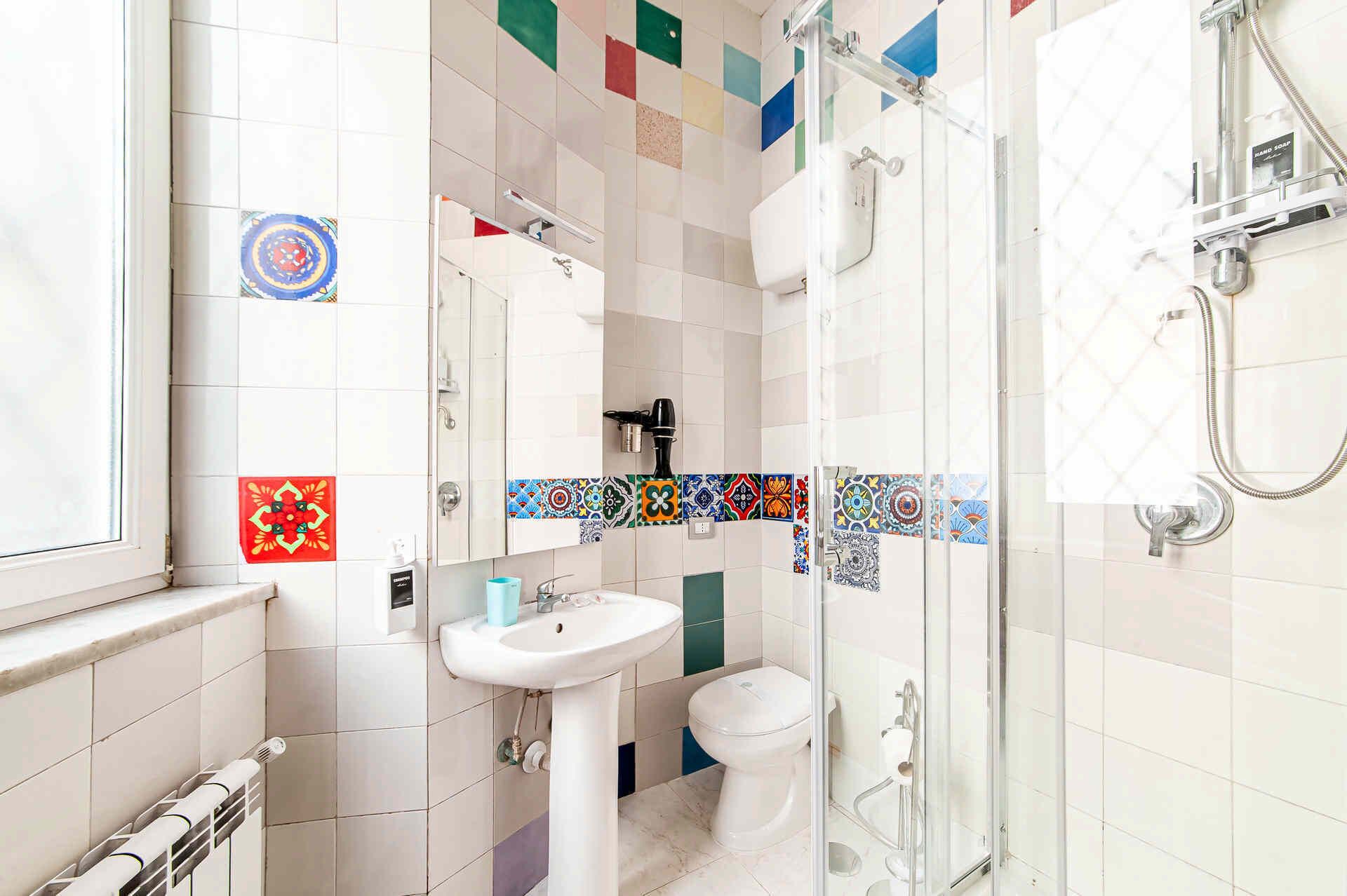 A bathroom with a white sink, toilet, and shower. The walls are tiled with colorful patterns.