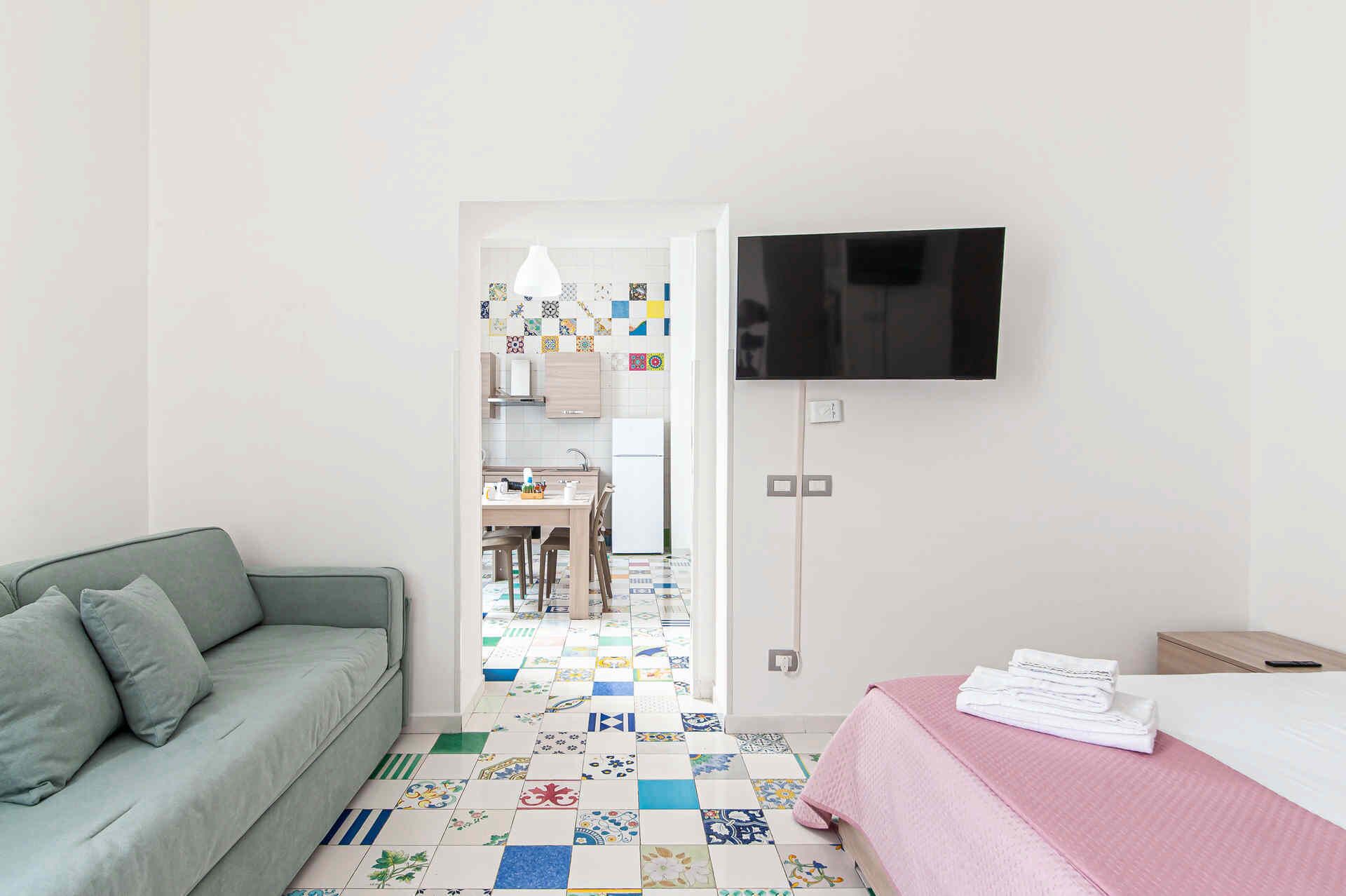 Une chambre d’hôtel avec un canapé gris, un lit recouvert d’une couverture rose, un téléviseur fixé au mur et un coin cuisine visible à travers une porte ouverte.