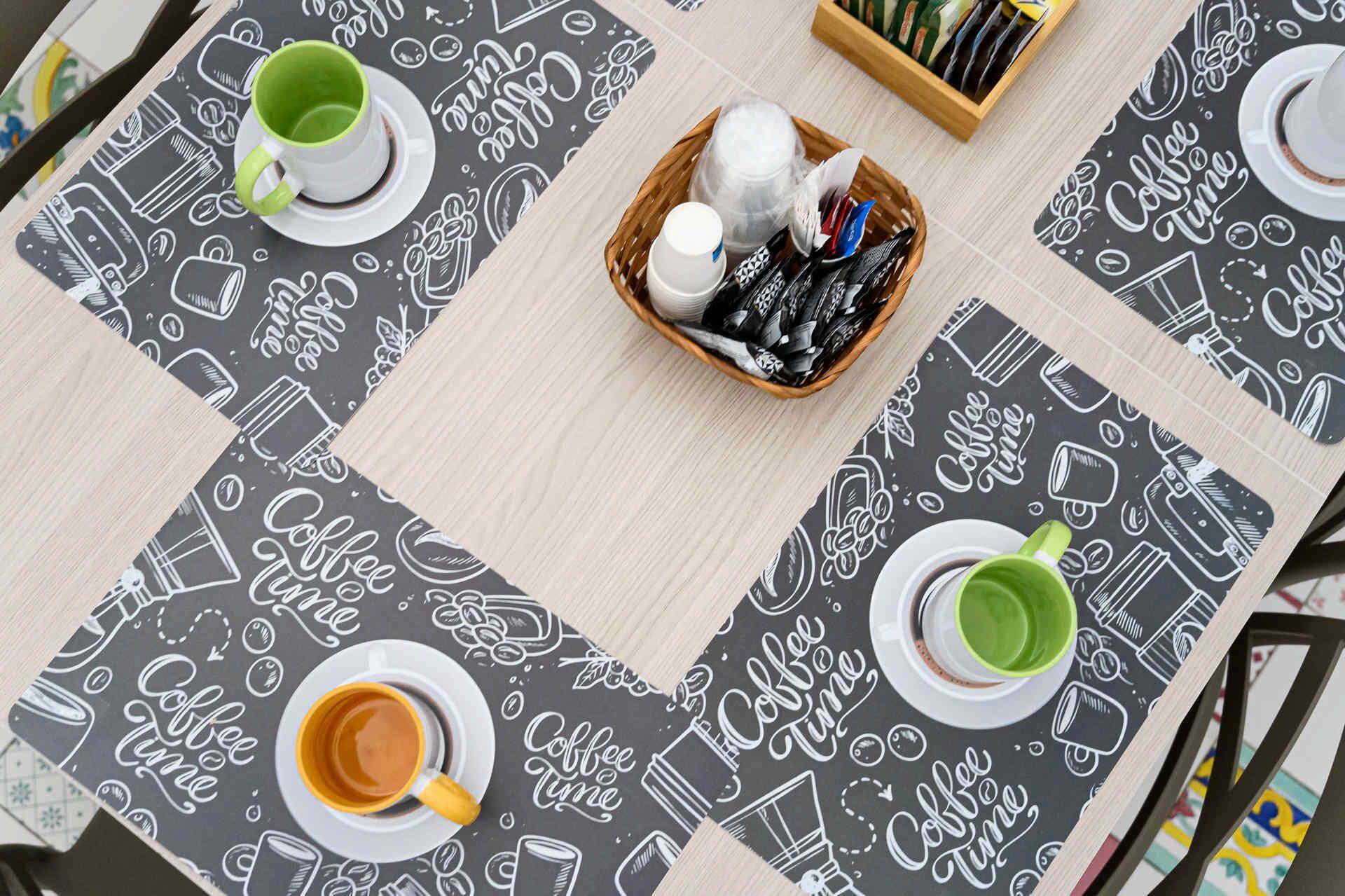 A table with a coffee-themed tablecloth, two cups of coffee, a basket with coffee-related items, and a coffee maker.
