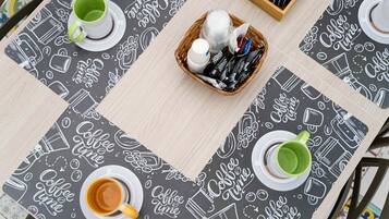 A table with a coffee-themed tablecloth, two cups of coffee, a basket with coffee-related items, and a coffee maker.