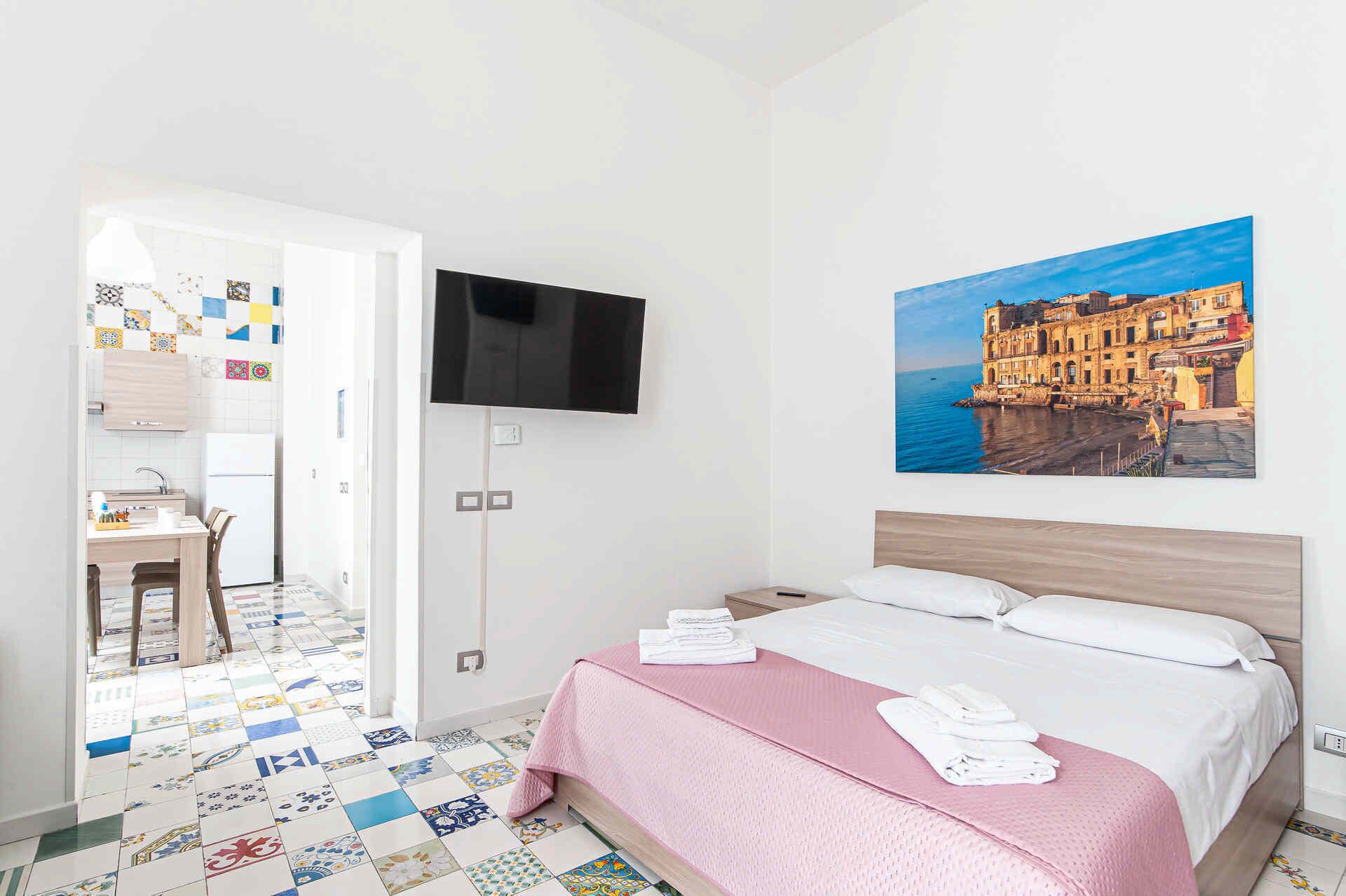 A hotel room with a bed, a TV, a painting of a coastal building, and a kitchen area with a tiled floor.