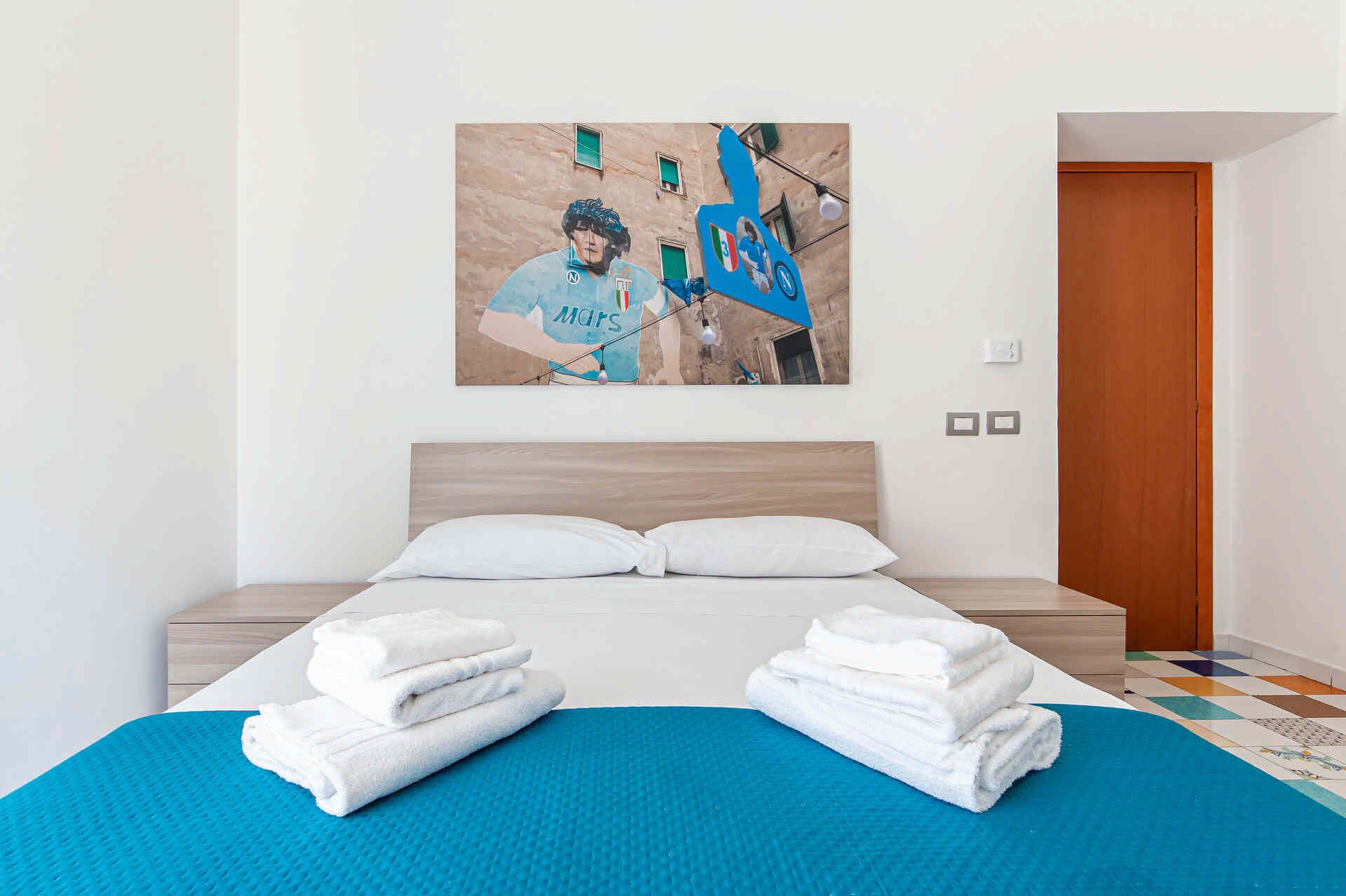 Quarto de hotel com uma cama, duas almofadas, um tapete azul e uma parede com uma pintura com tema esportivo.