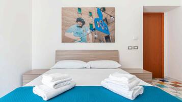 Quarto de hotel com uma cama, duas almofadas, um tapete azul e uma parede com uma pintura com tema esportivo.