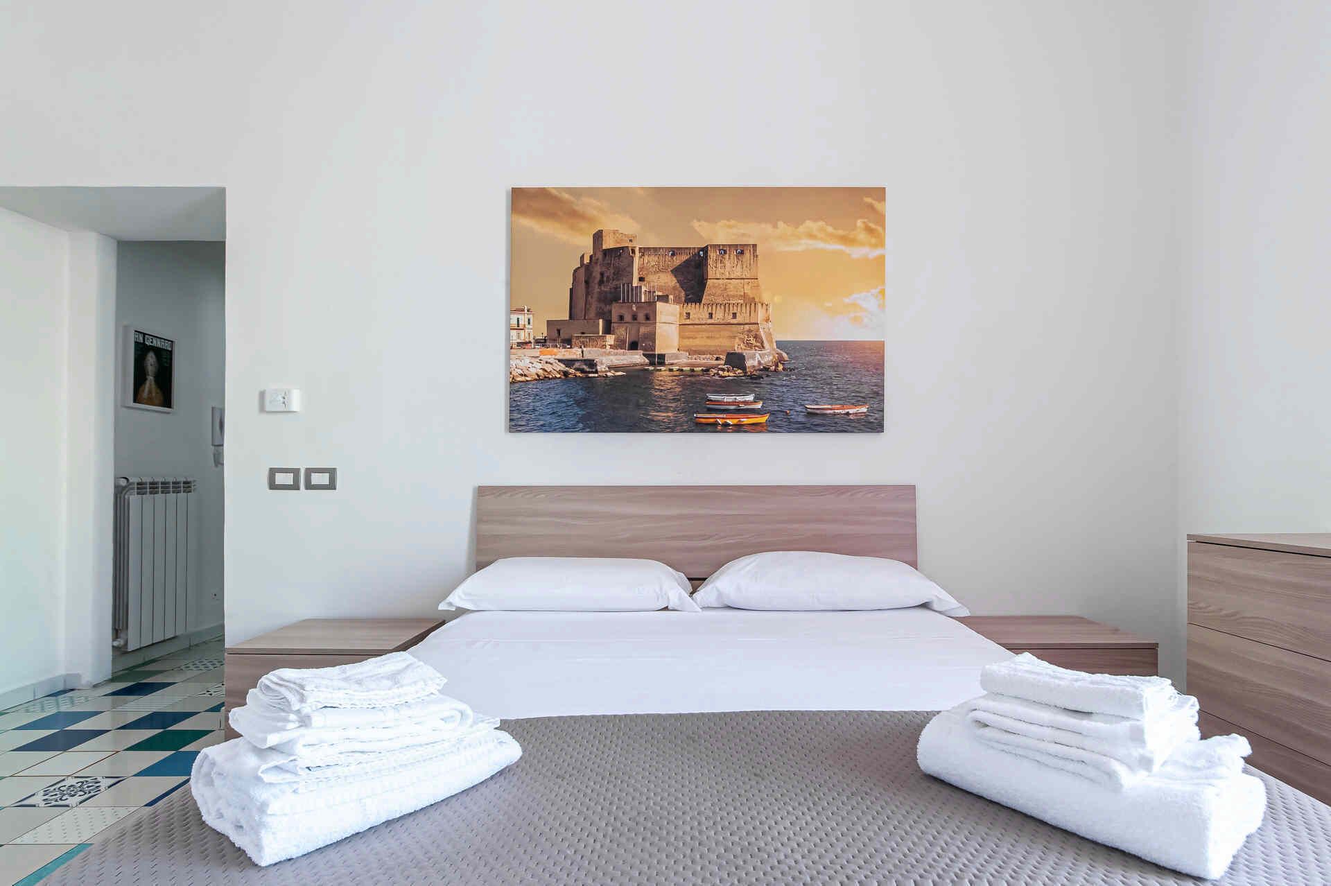 Camera d'albergo con un letto grande, due cuscini, una testiera, un comodino, un termosifone, un quadro appeso alla parete e vista su un castello sul mare.