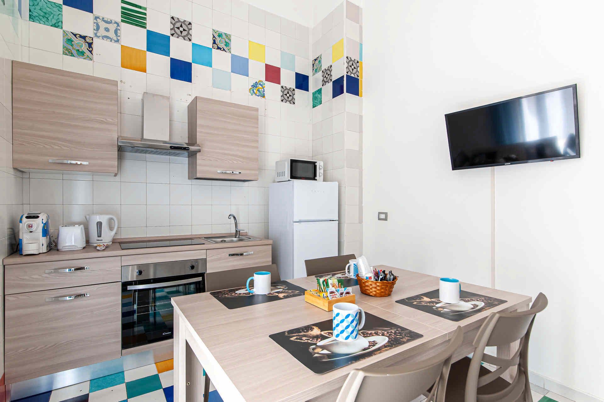 A modern kitchen with a microwave, toaster, and kettle, a dining table with placemats, and a flat-screen TV mounted on the wall.