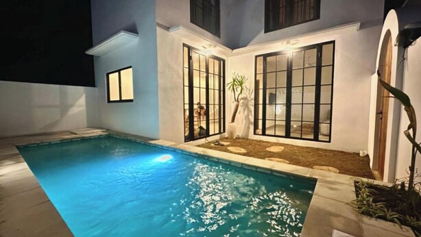 A swimming pool with a tiled deck, a small garden area with stepping stones, and a building with large windows and a white exterior. - Bukawa Residences Balangan 5 (Jimbaran)