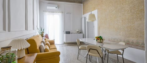 A living room with a sofa, a dining table with chairs, a kitchen area, and a patterned wall.