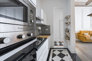 Een moderne keuken met een ingebouwde magnetron, oven en gasfornuis. Er ligt een geruit vloertapijt en er staat een gele fauteuil in de aangrenzende zithoek.