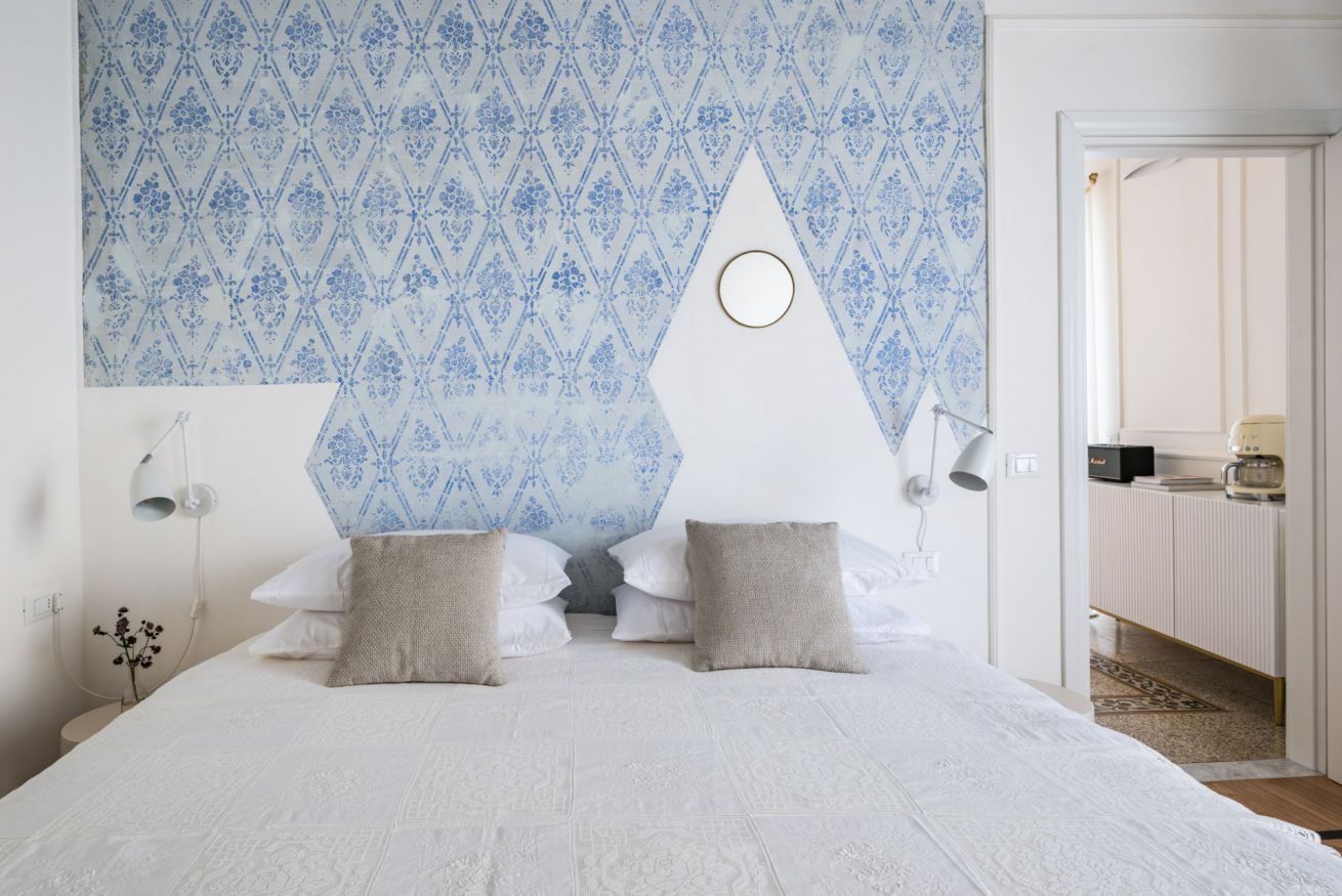 A bedroom with a patterned wallpaper, a bed with white bedding, two pillows, a bedside lamp, and a round wall decoration.