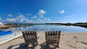 Una playa con dos sillas de madera mirando a un lago tranquilo, un kayak azul y una orilla rocosa con edificios al fondo.