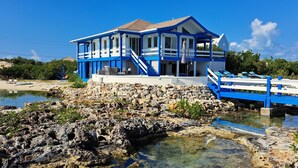Una casa frente a la playa, de color azul y blanco, con base de piedra, terraza de madera y vista despejada de la costa rocosa.