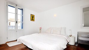 Ein Schlafzimmer mit Bett, Nachttisch, Lampe, kleinem Tisch mit Blumenvase, einem Schild an der Wand und einem Fenster mit Blick auf Gebäude.