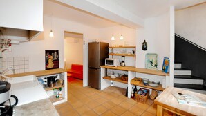 Eine kompakte Küche mit Kühlschrank, Mikrowelle und verschiedenen Küchenutensilien. Eine Holzauslage zeigt Geschirr, und ein kleiner Tisch ist mit Zeitschriften bestückt.