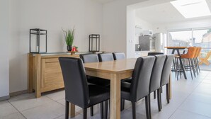 A modern dining area with a wooden table and black chairs, a kitchen in the background, and a large window providing natural light. - 4 bedroom gorgeous home in Petite-Synthe (Petite-Synthe)