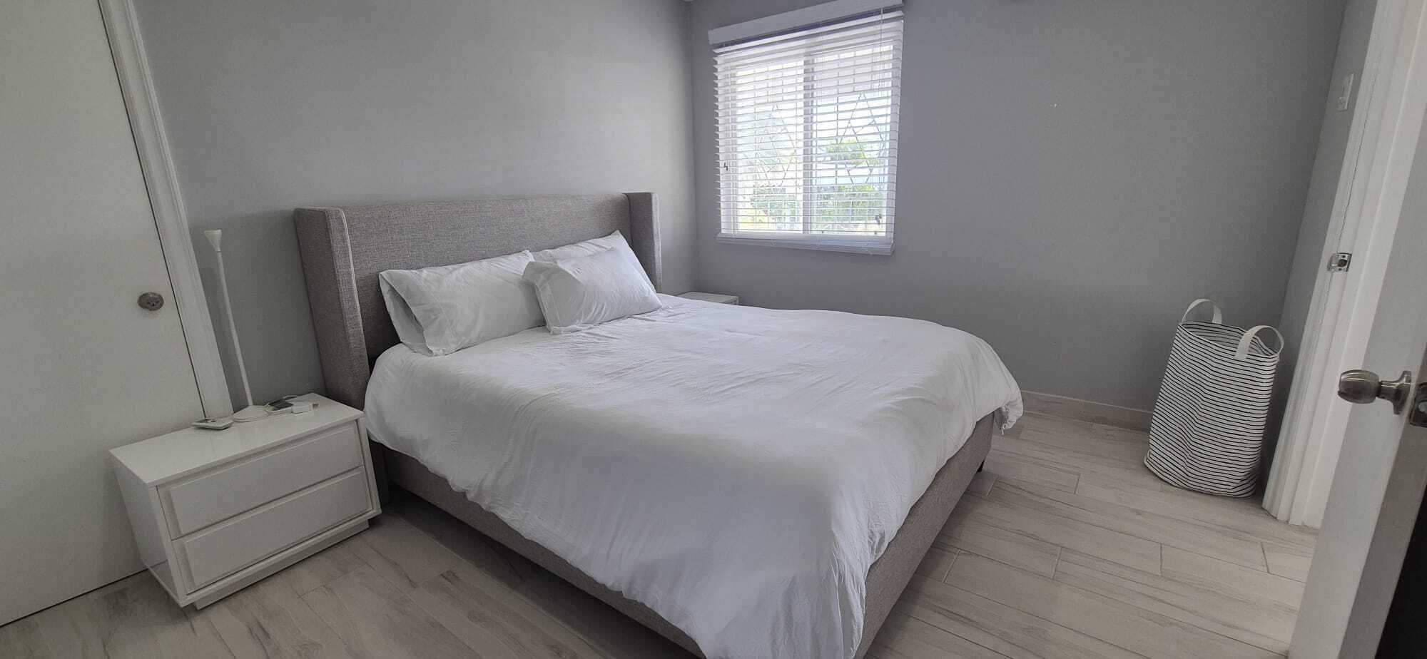 Une chambre à coucher avec un lit, une table de chevet, une fenêtre avec des stores et un sac à rayures près de la porte.