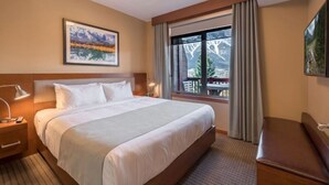 A hotel room with a large bed, a nightstand, a desk lamp, a window with a view of trees and mountains, and a painting on the wall.