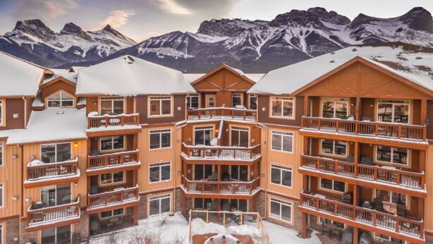 A multi-story lodge with balconies and a hot tub in front, set against a snowy mountain backdrop.
