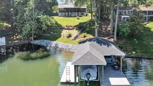 A houseboat docked on a calm lake, surrounded by lush greenery and a well-maintained lawn. - Pine and Paddle - Firepit / Water Toys / Gentle Lot / Foosball / Ping-Pong (Moneta)