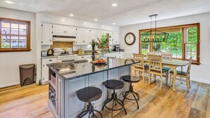 A spacious kitchen with a large island, white cabinetry, and a dining area with wooden chairs and a table. - Pine and Paddle - Firepit / Water Toys / Gentle Lot / Foosball / Ping-Pong (Moneta)