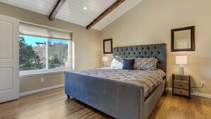 A bedroom with a large bed, wooden flooring, a window with blinds, and a ceiling with exposed beams. - Hot Tub, Fire Pit, Bocce Ball+ (Colorado Springs)
