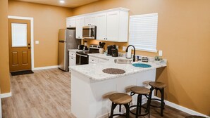 A compact kitchen with white cabinets, a refrigerator, a microwave, and a sink. There are stools at the island and a door leading to another room.