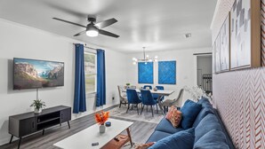 A modern living room with a sofa, coffee table, and TV. There is a dining area with chairs and a table. The room has a ceiling fan and artwork on the walls. - Spacious 4-bedroom Modern Bungalow in Charlotte! (Charlotte)
